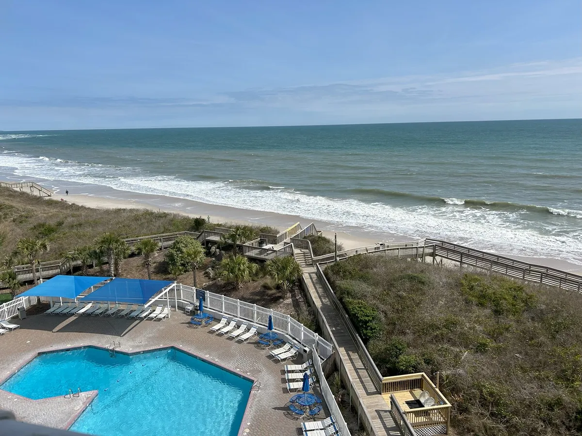 A wide exterior shot showing the resort’s beachside swimming pool, the ocean just beyond, and a wooden boardwalk leading to the sand. The inviting pool area is surrounded by lounge chairs and palm greenery, epitomizing the luxury amenities available to guests.