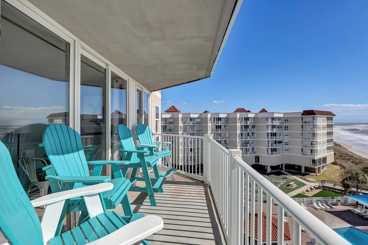 A pair of vibrant turquoise Adirondack chairs rest on a private balcony overlooking the sparkling ocean. Behind the chairs, modern resort buildings are visible, and the scene is bathed in soft, natural light, evoking a sense of tranquility and seclusion. The balcony is positioned to capture the beauty of sunrise or quiet moments shared by couples.