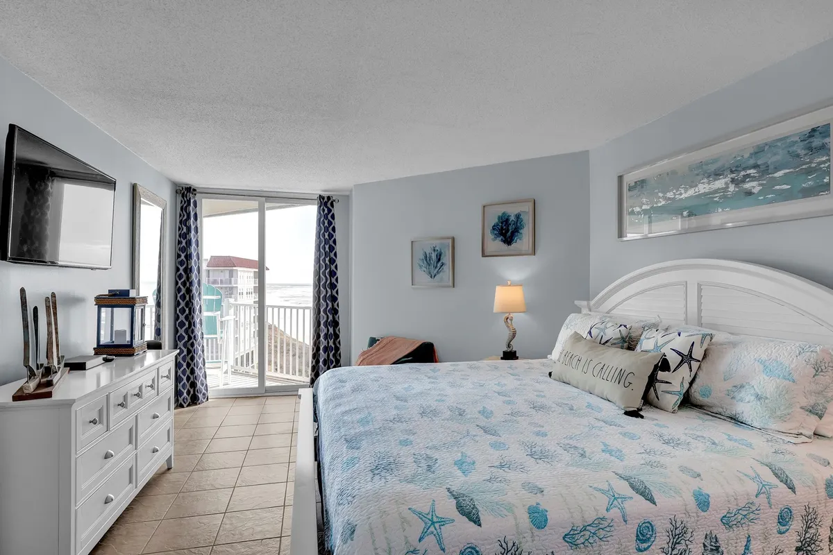 A spacious master bedroom featuring a king-sized bed adorned with beach-themed bedding, a large sliding glass door opening onto a balcony with ocean views, decorative artwork with marine motifs, and a wall-mounted television. Natural light floods the space, and the ocean can be glimpsed through the glass.