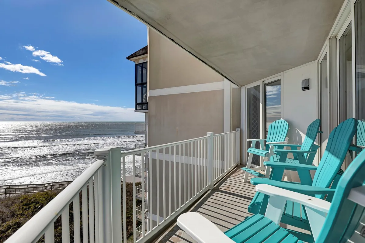 A tranquil balcony scene with adirondack chairs facing out over the ocean, separated by a simple balcony railing. The view extends across sparkling waves, inviting quiet mornings and golden sunsets.