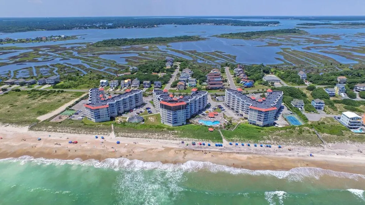 This image showcases a vibrant beachfront scene featuring condominium complexes set along the oceanfront, a sparkling swimming pool, marshland, and rolling ocean waves. The view captures the expansive outdoor amenities and the inviting connection to the beach, perfectly illustrating the getaway’s coastal atmosphere.
