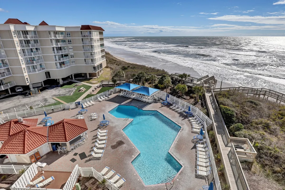 The image showcases a stunning oceanfront condo complex with a sparkling, brand new saltwater covered pool at the center. Lounge chairs line the poolside, inviting relaxation, while the expansive ocean stretches beyond, framed by the modern building structure. The tranquil setting is enhanced by soft sunlight, evoking a sense of both sunrise serenity and the promise of beautiful sunset views.