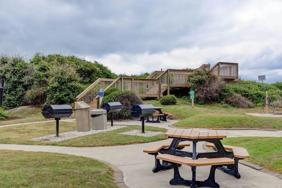 This image features a communal outdoor area with picnic tables, grill stations, and a wooden stairway leading toward the beach. The scene is lively and communal, suggesting group barbecues, shared meals, and time spent socializing outdoors after a day of adventure.