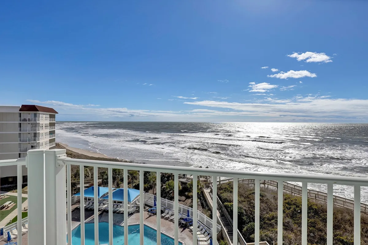 A sweeping view from the balcony highlights the sparkling ocean, the shimmering swimming pool below, and the sturdy balcony railing in the foreground. The scene is bathed in natural light, with tranquil blue tones of the sea and sky, inviting viewers to imagine early mornings or evenings spent enjoying the serenity.
