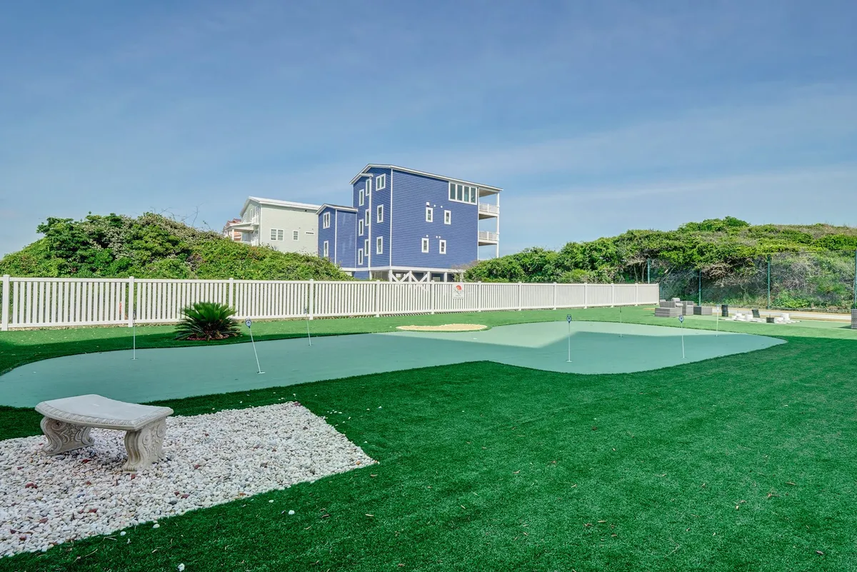 A picturesque putting green with a decorative stone bench and lush landscaping, situated beside a blue house. The area is designed for friendly golf competitions, casual gatherings, or relaxing outdoors, and evokes the playful, social spirit of the amenities listed in the section.