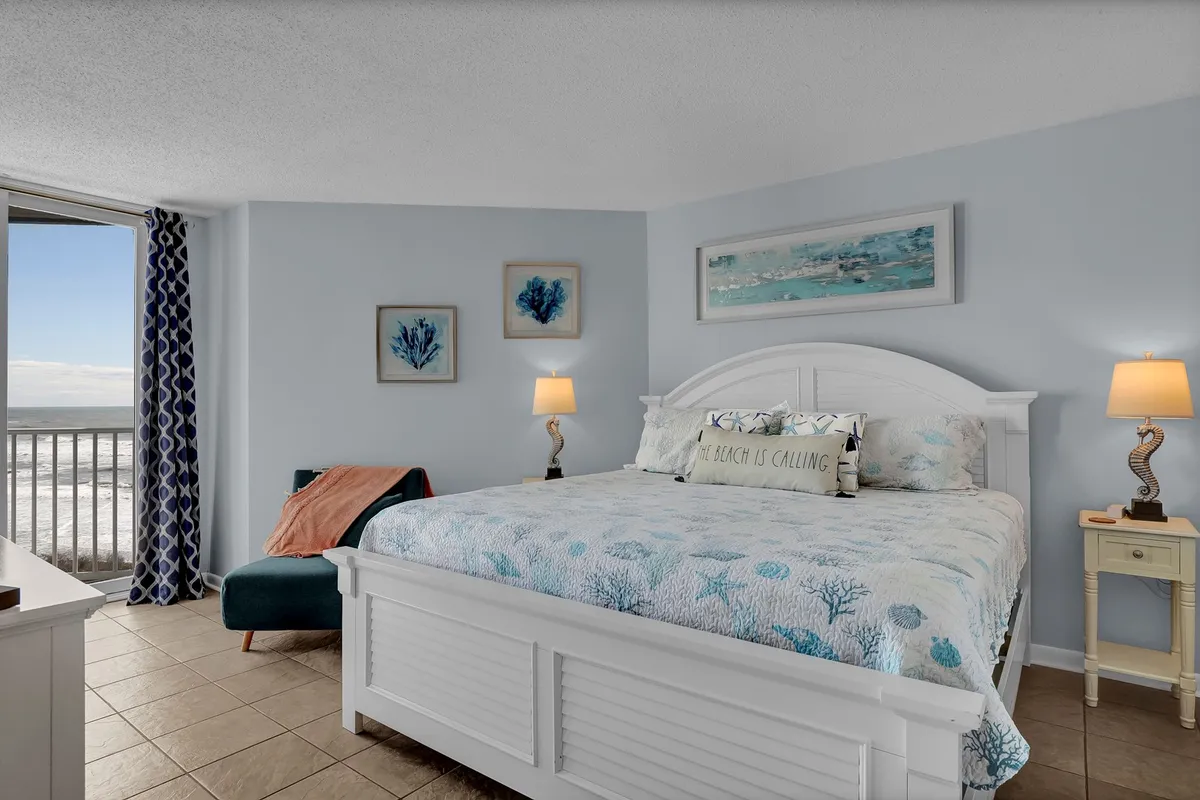 A tranquil bedroom scene showcasing a bed with blue coral-patterned bedding positioned beside a large window offering an ocean view. The room is decorated with coastal wall art and bedside lamps, radiating a serene, personalized retreat feel.