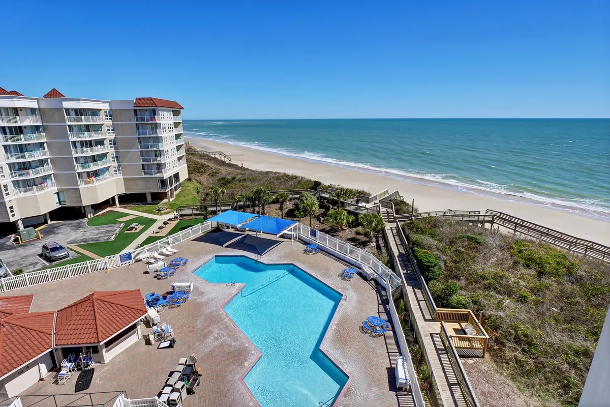 A stunning view of a sparkling swimming pool surrounded by greenery, with the beach and ocean in the background. The pool area features lounge chairs and is connected visually to the beach, suggesting seamless access between resort amenities and natural surroundings.