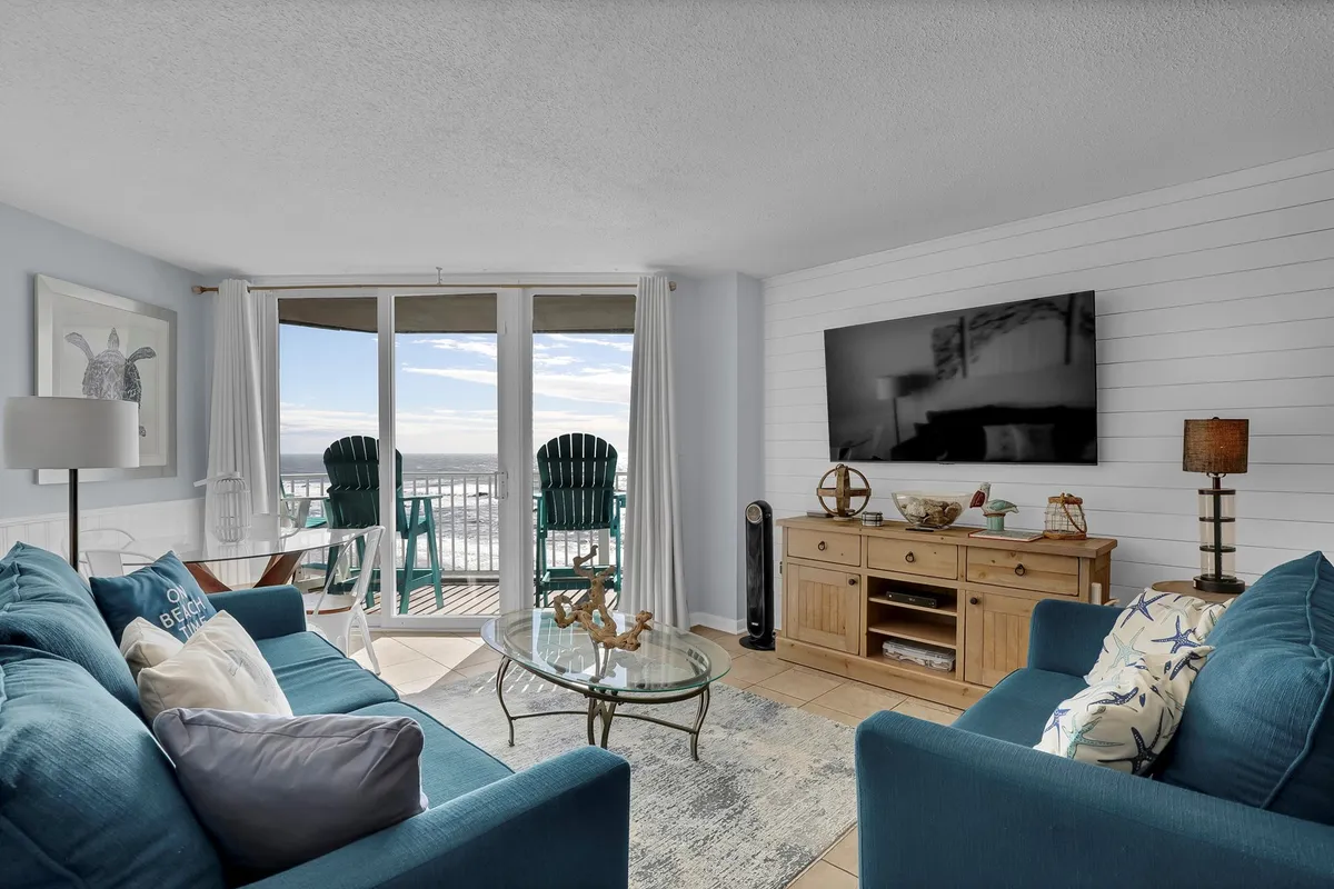 A bright, inviting living room with comfortable blue sofas facing large sliding glass doors that open to a private balcony with expansive ocean views. Wooden furniture accentuates the coastal decor, and sunlight fills the space, blurring the boundary between indoors and the Atlantic horizon.